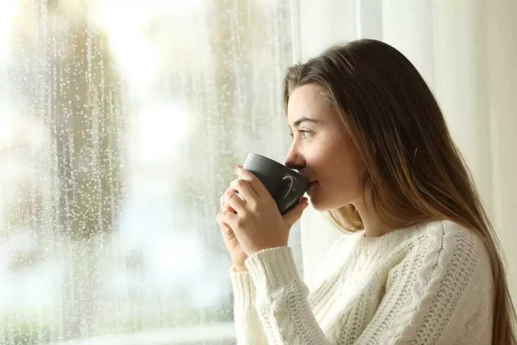 A young woman drinking tea and looking out a window at the rain.